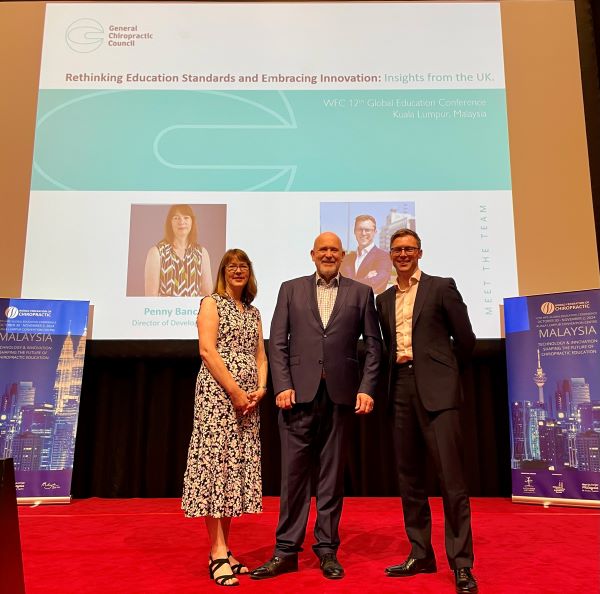 (left to right) Penny Bance (GCC), Richard Brown (World Federation of Chiropractic), Danile Moore (GCC) on the stage at the conference in front of the title slide to their keynote speech. 