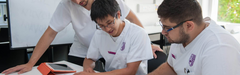 Three students studying together at a chiropractic university, reviewing textbooks and notes in a classroom setting.