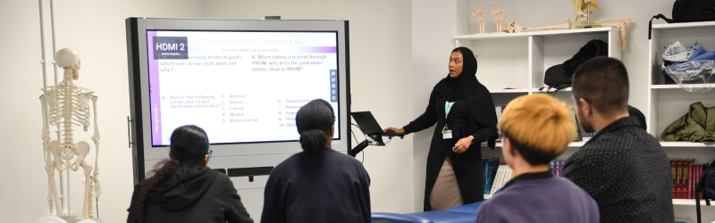 Teacher presenting a lecture on a screen to chiropractic students in a classroom, with a skeleton model positioned on the left side of the room.