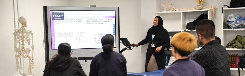 Teacher presenting a lecture on a screen to chiropractic students in a classroom, with a skeleton model positioned on the left side of the room.